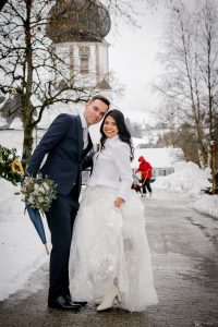 Brautpaar bei Brautpaarshooting mit der Fotografin Isabela Campos. Sie stehen im Schnee und schauen einander an. Die Braut hält das Gesicht des Bräutigams. Im Hintergrund ist die Kirche von Kirchzarten zu sehen.