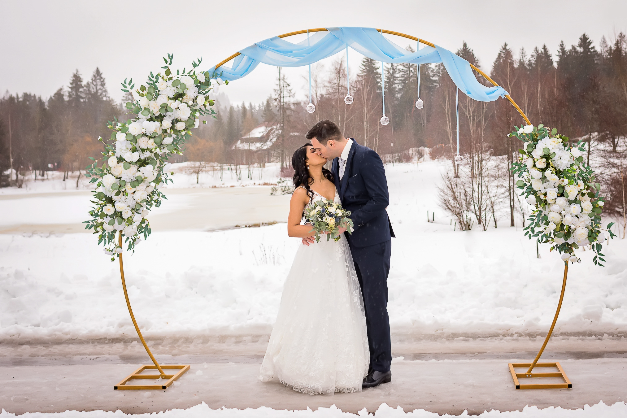 Ein Brautpaar steht in einer wunderschönen Winterlandschaft des Schwarzwaldes in Hinterzarten vor einem mit weißen Blumen dekorierten Hochzeitsbogen und umarmt und küsst sich leidenschaftlich. Fotografiert von der talentierten Fotografin Isabela Campos.
