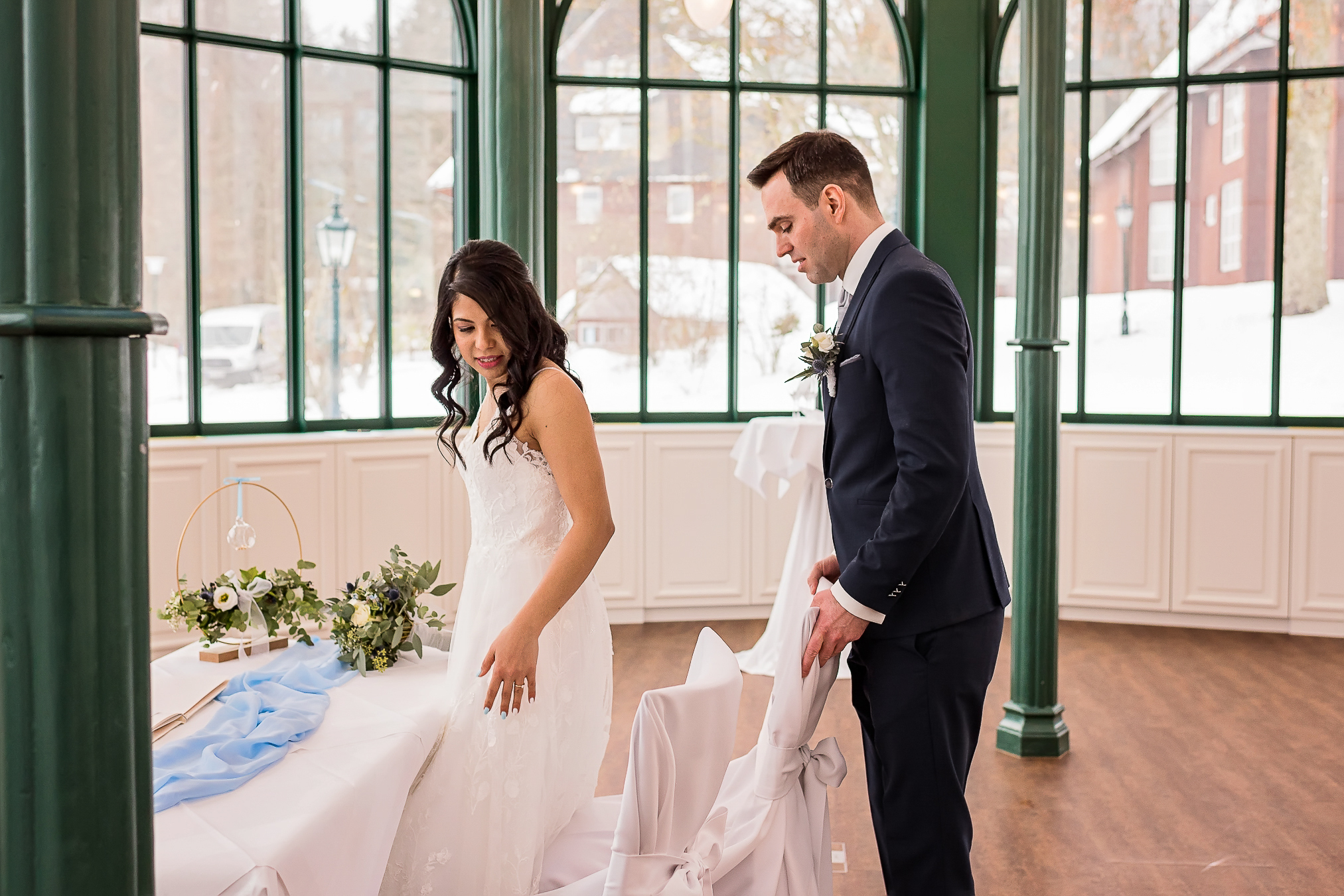 Brautpaar betritt Pavillon Diva im verschneiten Parkhotel Adler in Hinterzarten. Der Bräutigam hilft der Braut, sich hinzusetzen, während sie in ihre wunderschöne Winterhochzeitskulisse schauen.