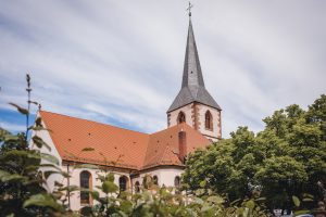 Ein Foto der beeindruckenden Fassade der Evangelischen Kirche in Friesenheim während einer kirchlichen Hochzeit. Der Himmel ist blau und es scheint ein strahlender Sonnentag zu sein. Der Bildausschnitt zeigt einen Teil des Kirchendachs und des Turms, der majestätisch in den Himmel ragt.