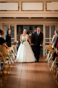 Ein Foto, das den bewegenden Moment zeigt, in dem die Braut von ihrem Vater in die Evangelische Kirche in Friesenheim geführt wird. Das Bild zeigt eine Nahaufnahme der Braut und ihres Vaters, die Hände halten, während sie die Kirche betreten.