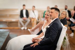 Ein Foto vom Brautpaar, das bei einer kirchlichen Hochzeit einem Auftritt eines Sängers oder einer Sängerin zusieht.