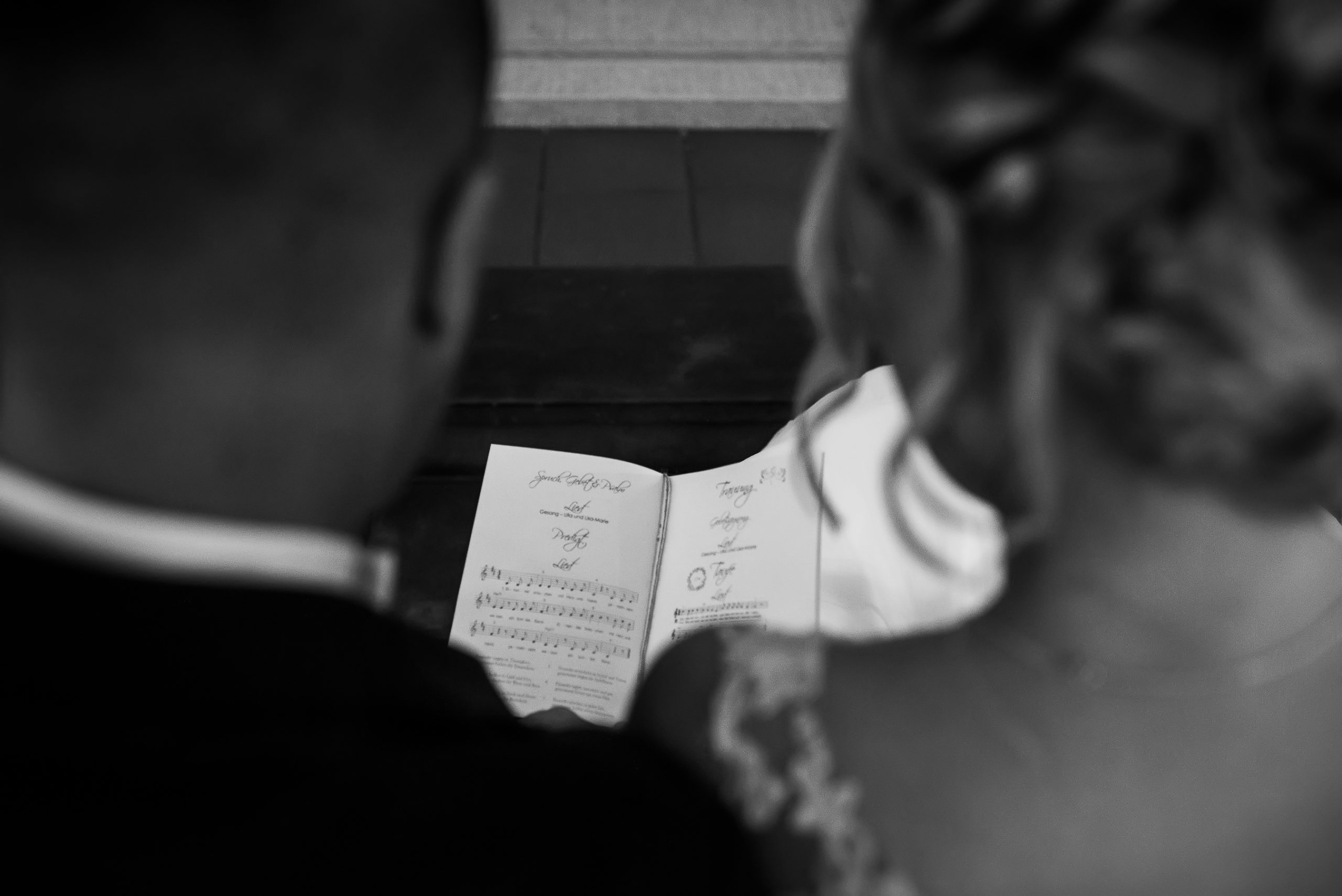 Ein schwarz-weiß Foto von einer romantischen kirchlichen Hochzeit in Offenburg. Das Brautpaar sitzt in der Kirche und schaut gemeinsam auf das Liederheft. Die Kamera ist von oben auf das Paar gerichtet und fängt das Detail ein.