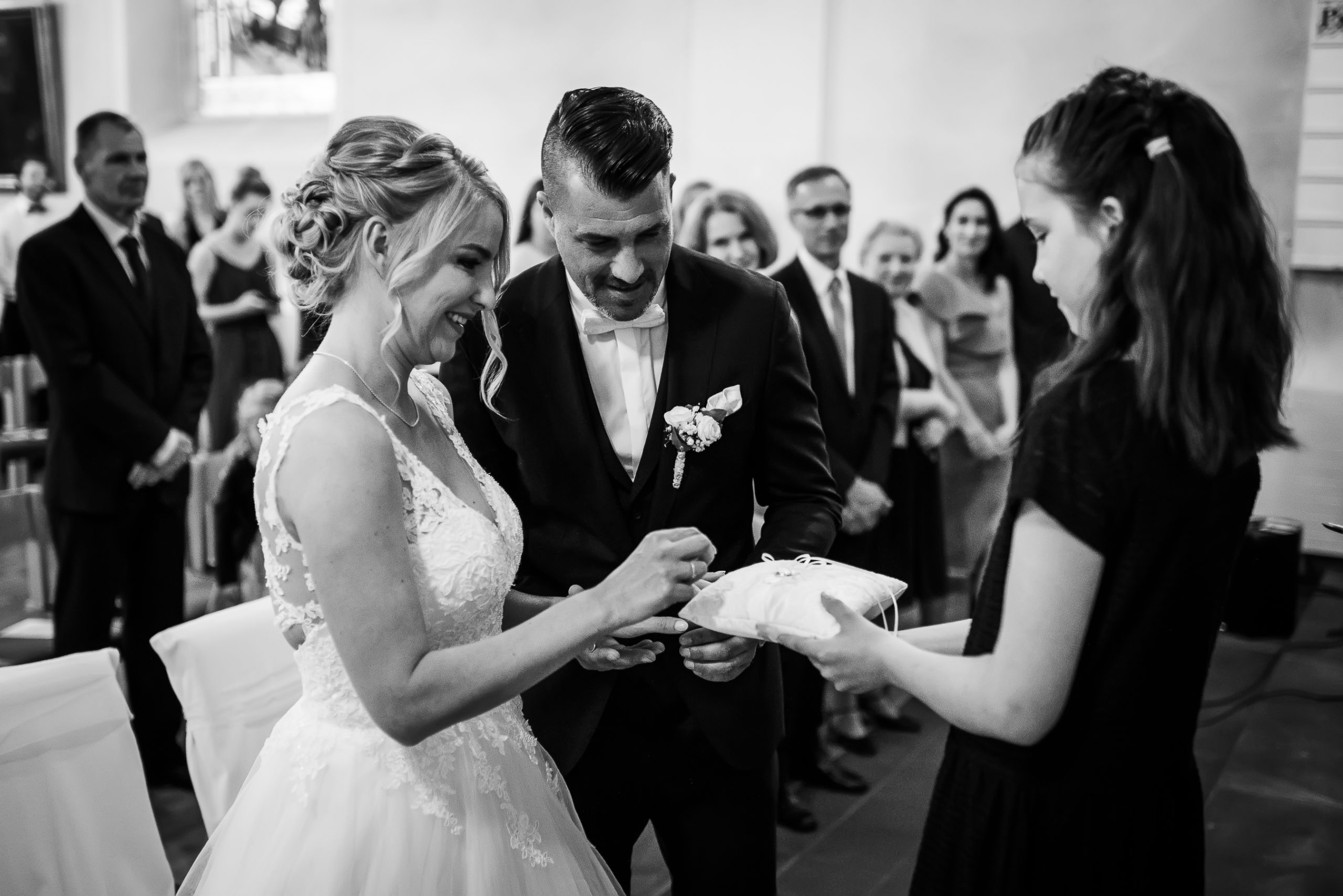 Schwarz-weiß-Foto von der ringtauschenden Zeremonie bei einer kirchlichen Hochzeit in Friesenheim. Die Braut steckt den Ring auf die Hand des Bräutigams, während die Tochter das Hochzeitskissen hält.