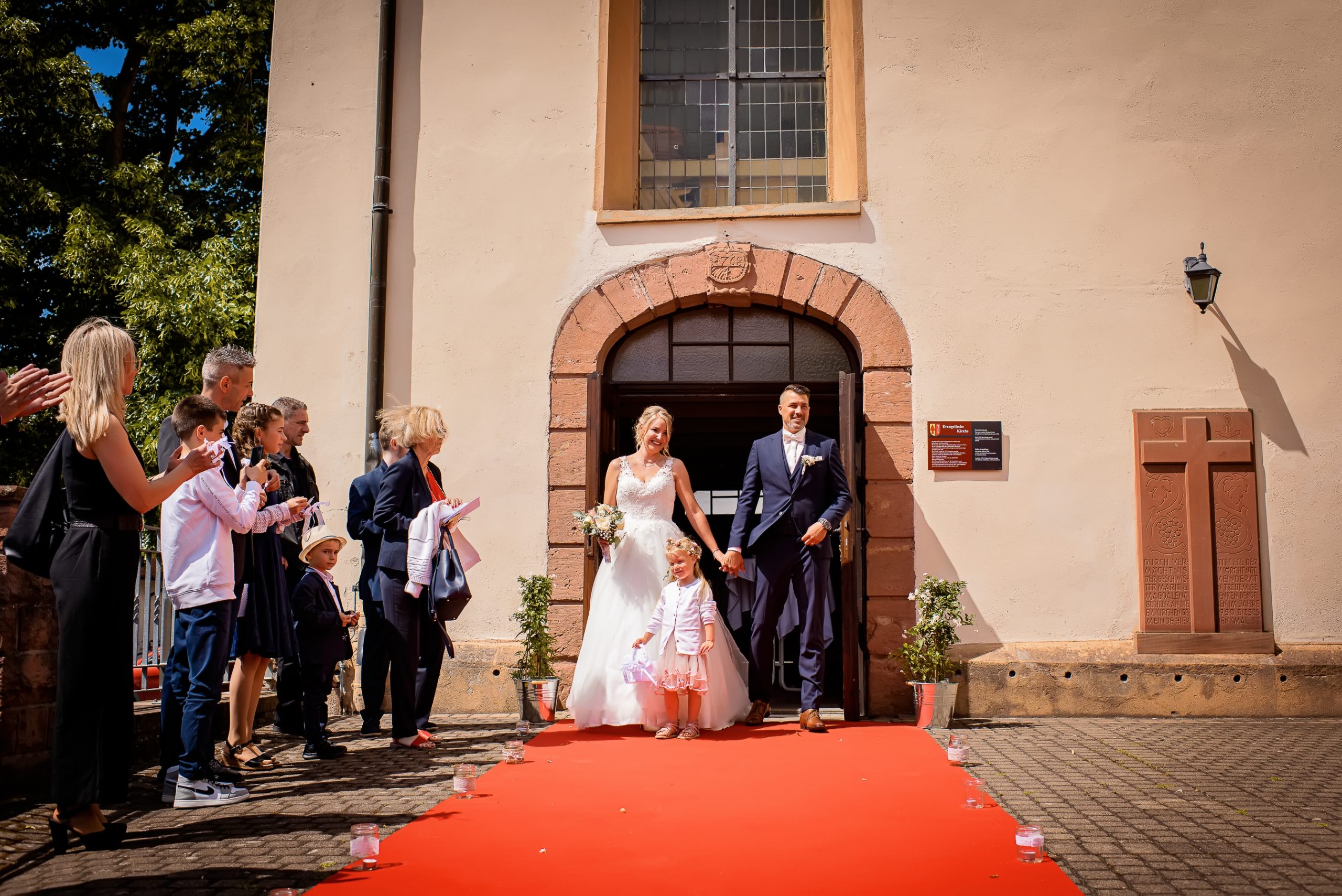 Ein glückliches Brautpaar verlässt die Evangelische Kirche in Friesenheim an einem sonnigen Sommertag, nachdem sie gerade ihre Hochzeitszeremonie abgeschlossen haben. Vor der Kirche liegt ein roter Teppich ausgerollt, der den Weg in eine neue gemeinsame Zukunft symbolisiert.