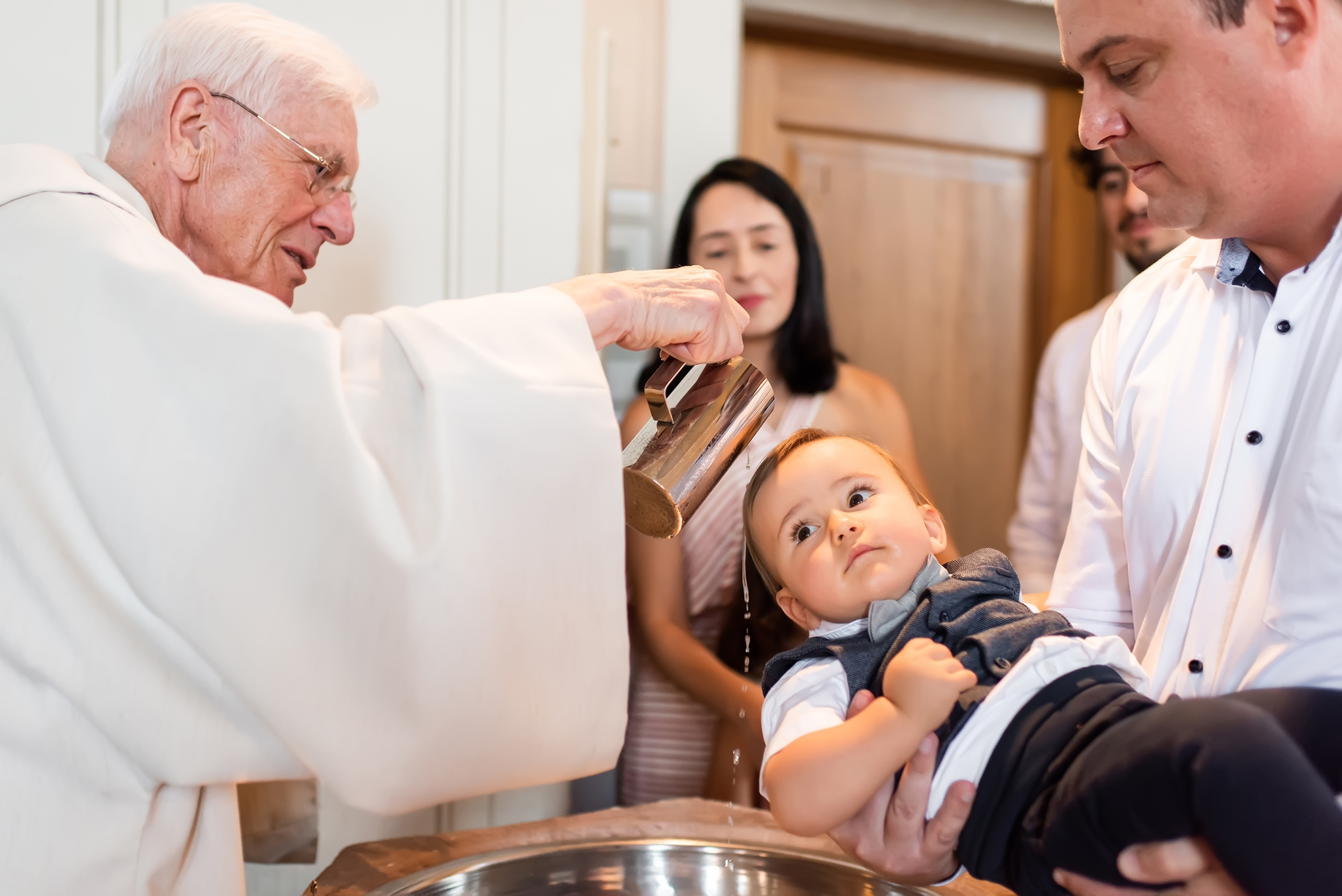Ein Foto einer Taufzeremonie in Durbach, bei der das Kind vom Pfarrer oder Geistlichen mit heiligem Wasser getauft wird. Das Foto zeigt im Detail das Taufritual.