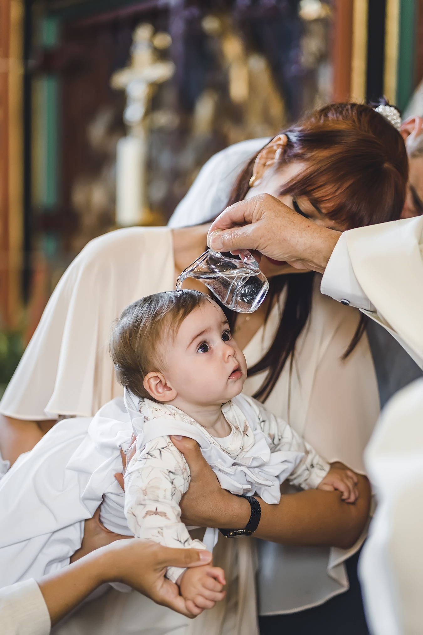 Ein Foto einer Taufzeremonie in Ehrenkirchen bei Freiburg, bei der das Pfarrer das Kind mit heiligem Wasser tauft. Das Foto zeigt im Detail das Taufritual bei einem Mädchen. Die Tante hält das Baby im Arm und das Baby schaut neugierig zum Pfarrer.