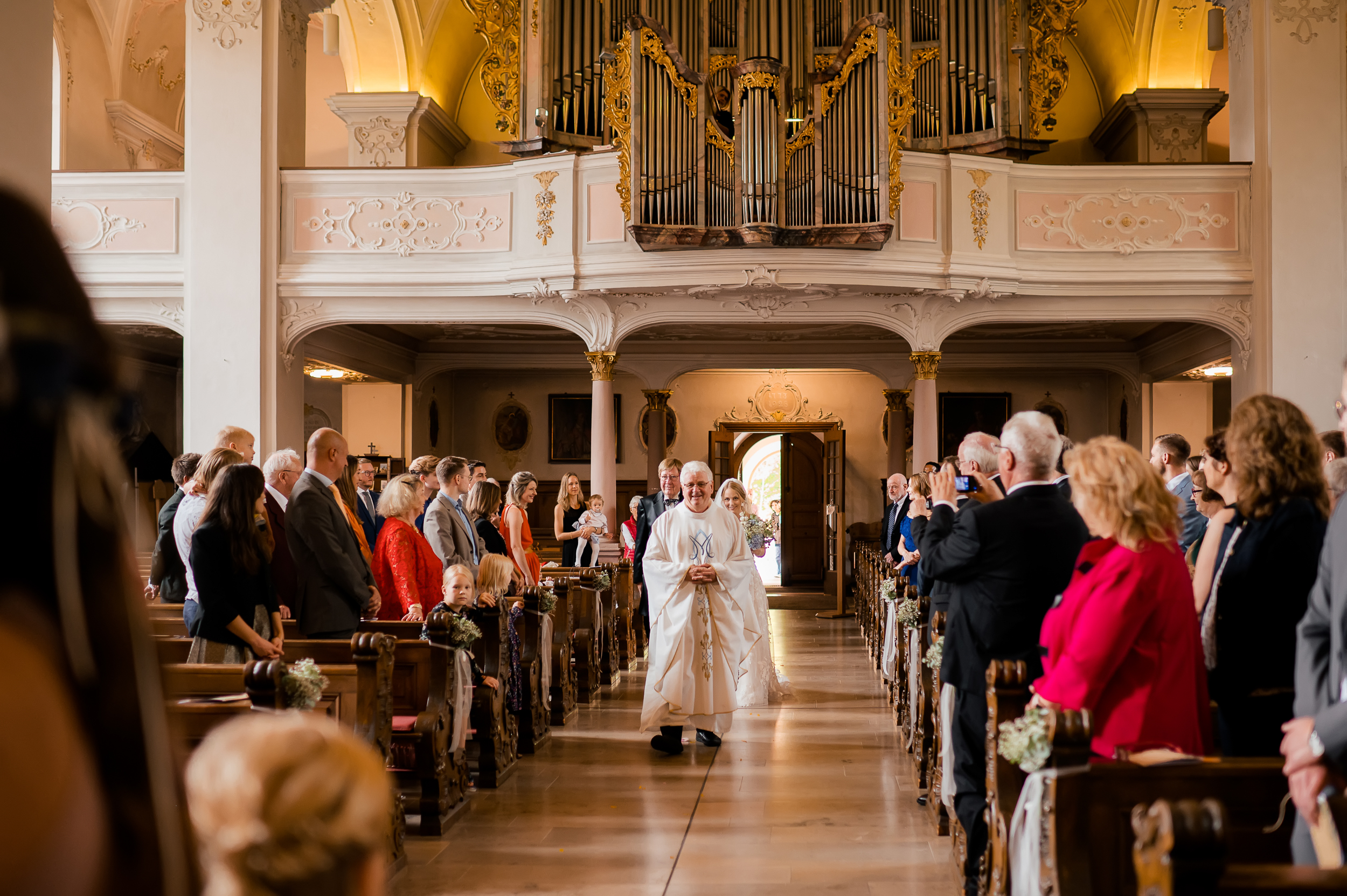 Pater Roman in der Wallfahrtskirche Todtmoos läuft zum Altar und hinter ihm sind die Braut und ihr Vater. Die Hochzeitsgäste stehen und schauen die Braut an.