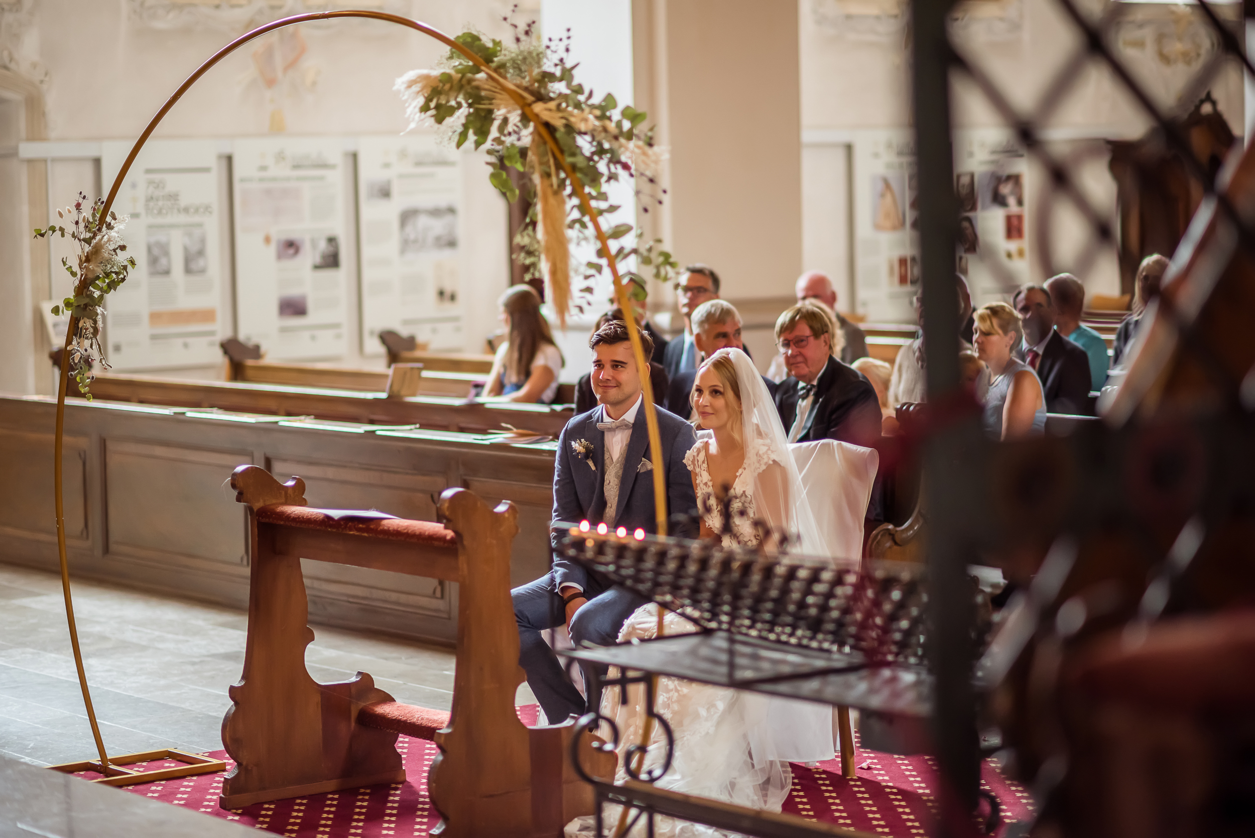 Brautpaar sitzt vor einem goldenen Hochzeitsbogen im Altar einer katholischen Kirche, die Braut trägt einen weißen Schleier.