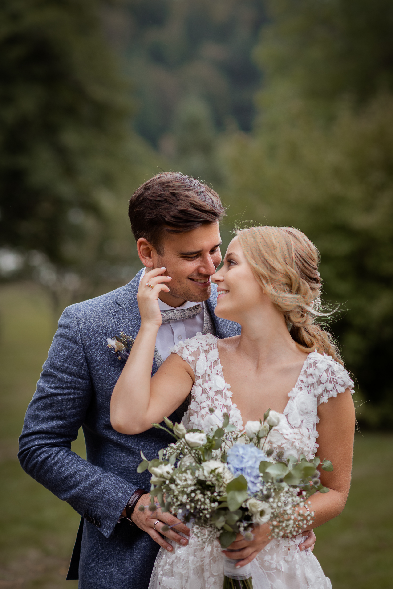 Ein Brautpaar Fotoshooting im Kurpark in Todtmoos. Der Bräutigam umarmt die Braut von hinten, während sie sich in die Augen schauen. Die Braut streicht sanft über das Gesicht des Bräutigams und hält in der linken Hand ihren Blumenstrauß.