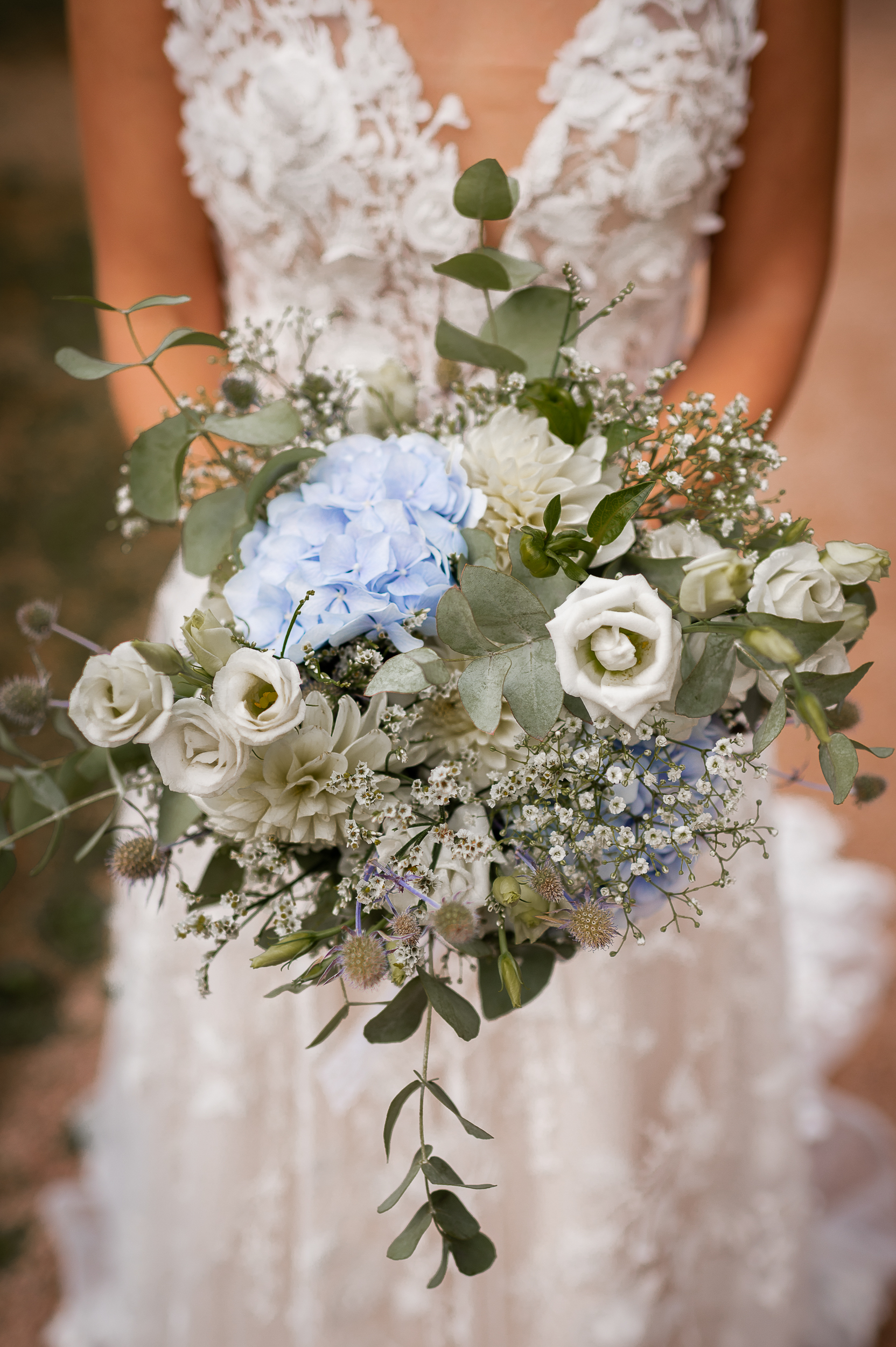 Nahaufnahme von einem Brautstrauß im Boho-Stil mit weißen Minirosen und hellblauen Hortensien, unscharf im Hintergrund ist das Spitzenkleid der Braut zu sehen.