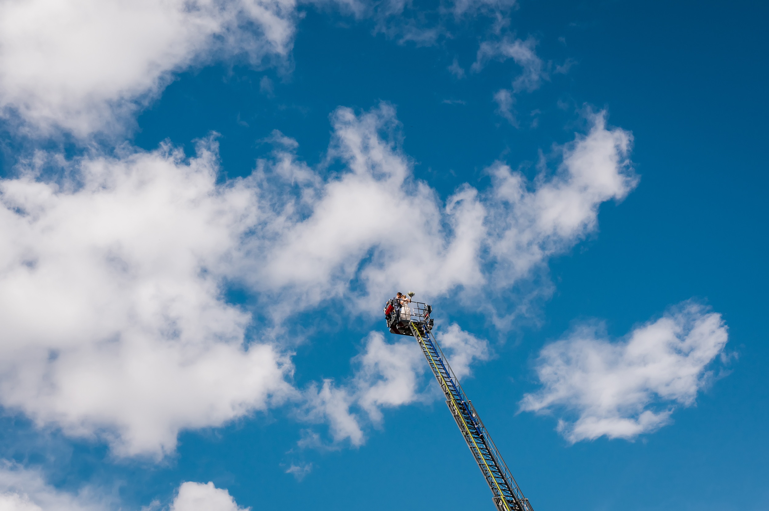 Ein Brautpaar wird auf einem Feuerwehrauto in die Luft gehoben und küsst sich inmitten eines wunderschönen blauen Himmels.