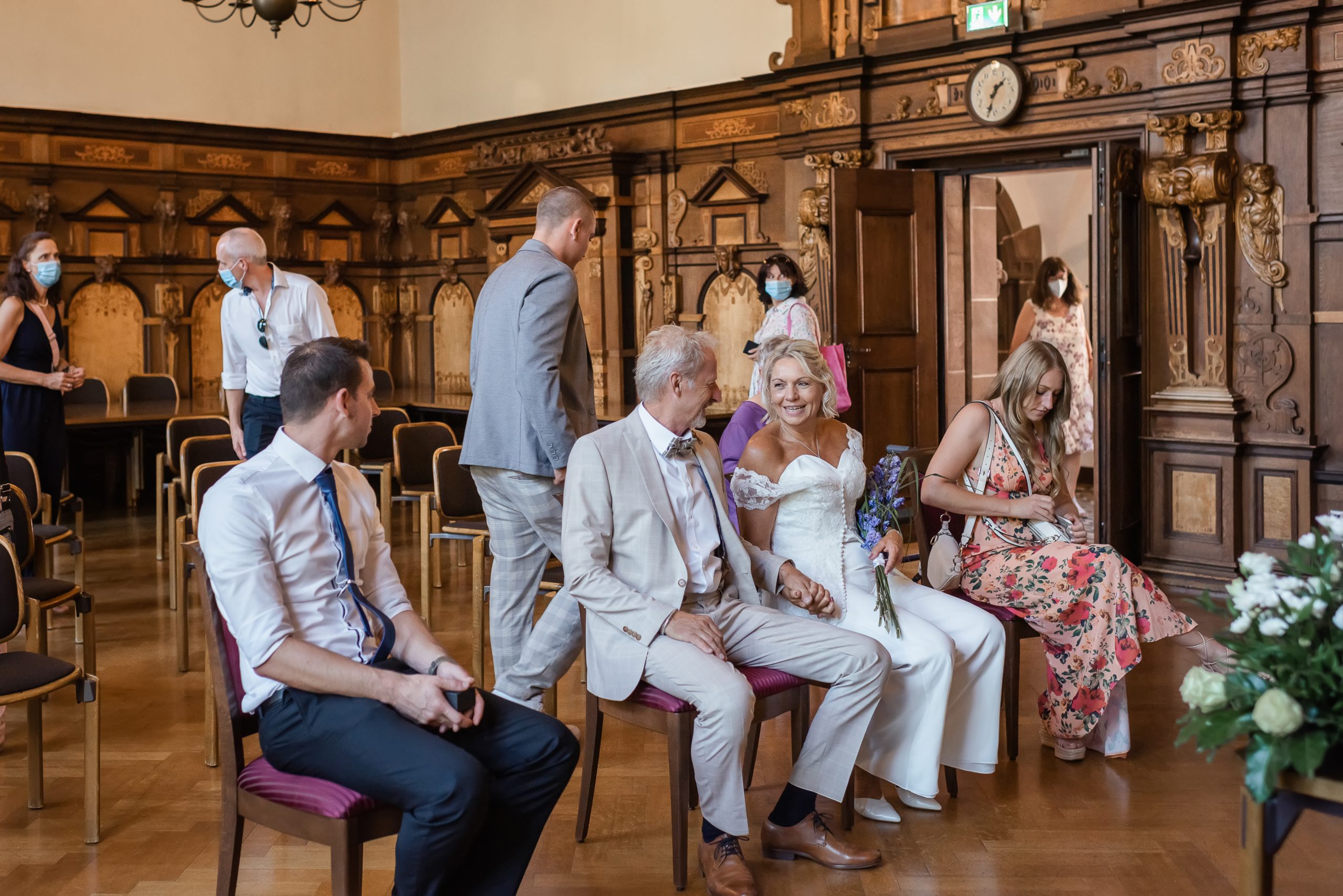 Brautpaar hält Hand in Hand im Trausaal des Freiburger Rathauses, umgeben von ihren Gästen, während sie auf den Beginn ihrer standesamtlichen Zeremonie warten.