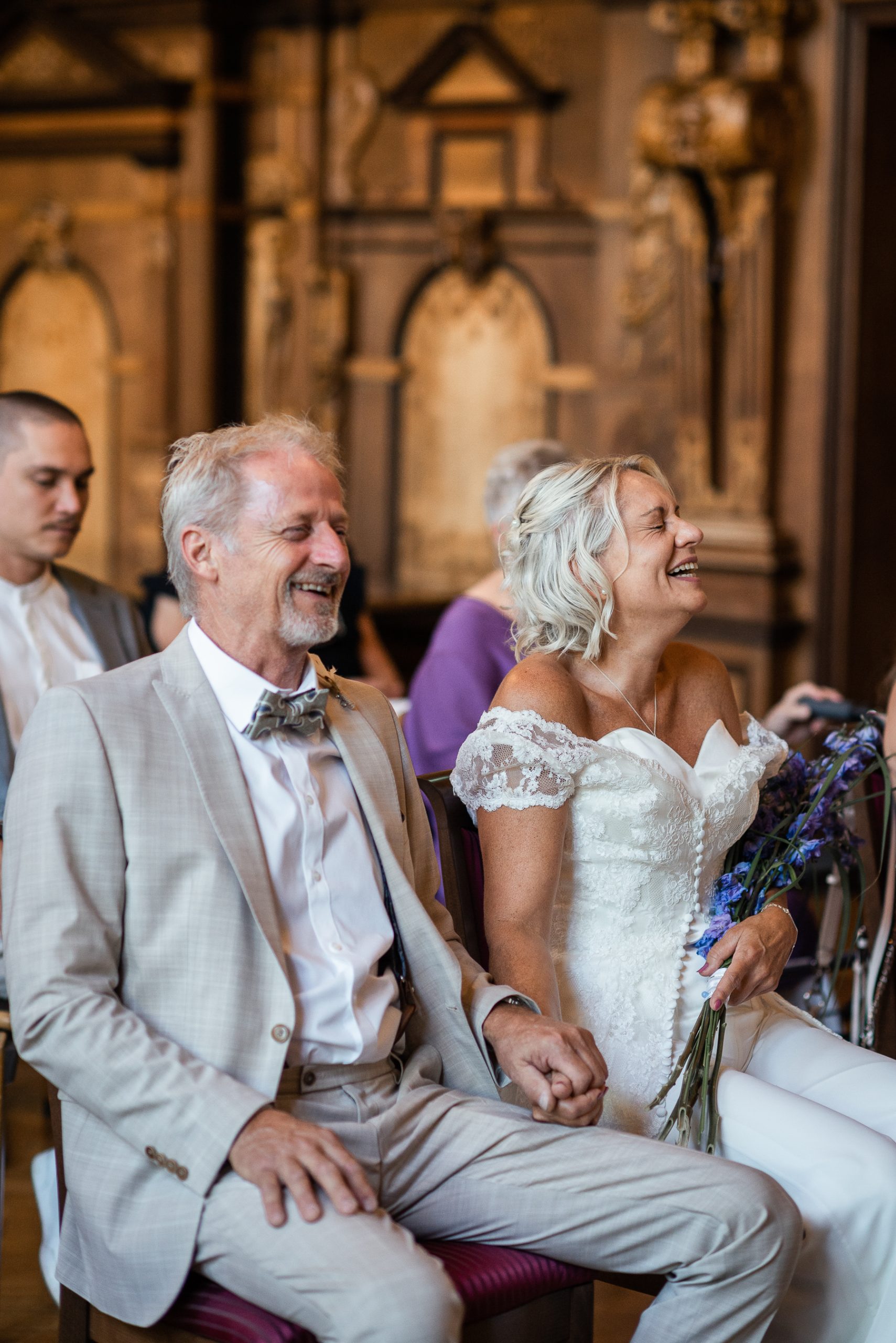Ein Brautpaar sitzt Hand in Hand bei einer standesamtlichen Zeremonie im Trausaal des Freiburger Rathauses und lacht fröhlich.