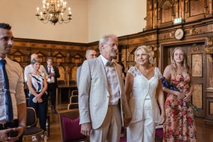 Ein glückliches Brautpaar hält Hand in Hand im Trausaal des Freiburger Rathauses und gibt sich das Ja-Wort bei ihrer standesamtlichen Zeremonie in Freiburg, während sie von ihren Gästen umgeben sind. Die Fotografin Isabela Campos hält das emotionale Ereignis fest.