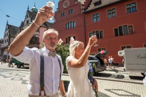Ein Brautpaar und ihre Gäste stoßen mit Aperitifs vor dem Alten Rathaus in Freiburg auf die Hochzeit an.