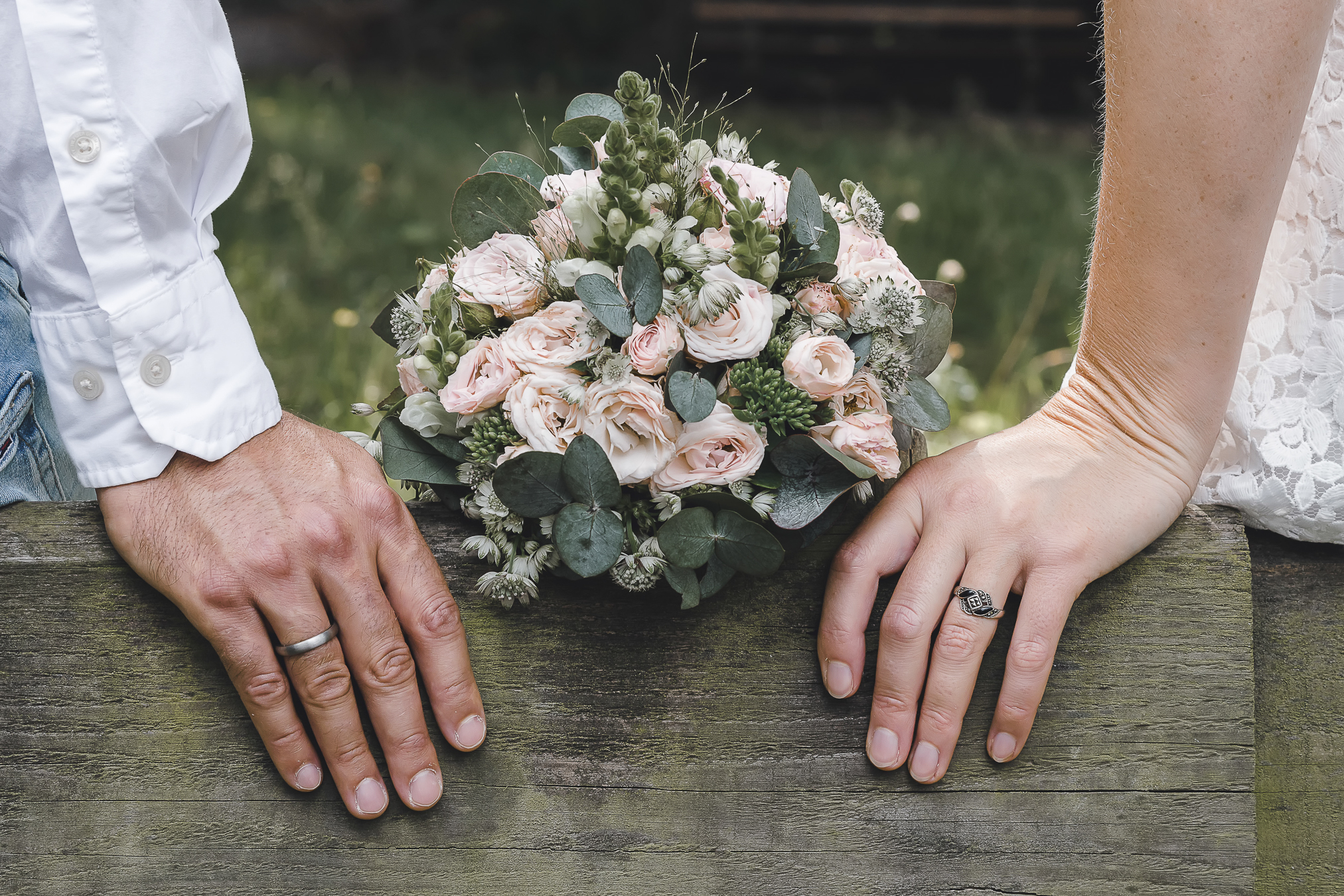 Eine Nahaufnahme der Hände des Brautpaars, wie sie sich an einem rustikalen Holzstamm anlehnen, während ein bezaubernder Blumenstrauß in der Mitte liegt.