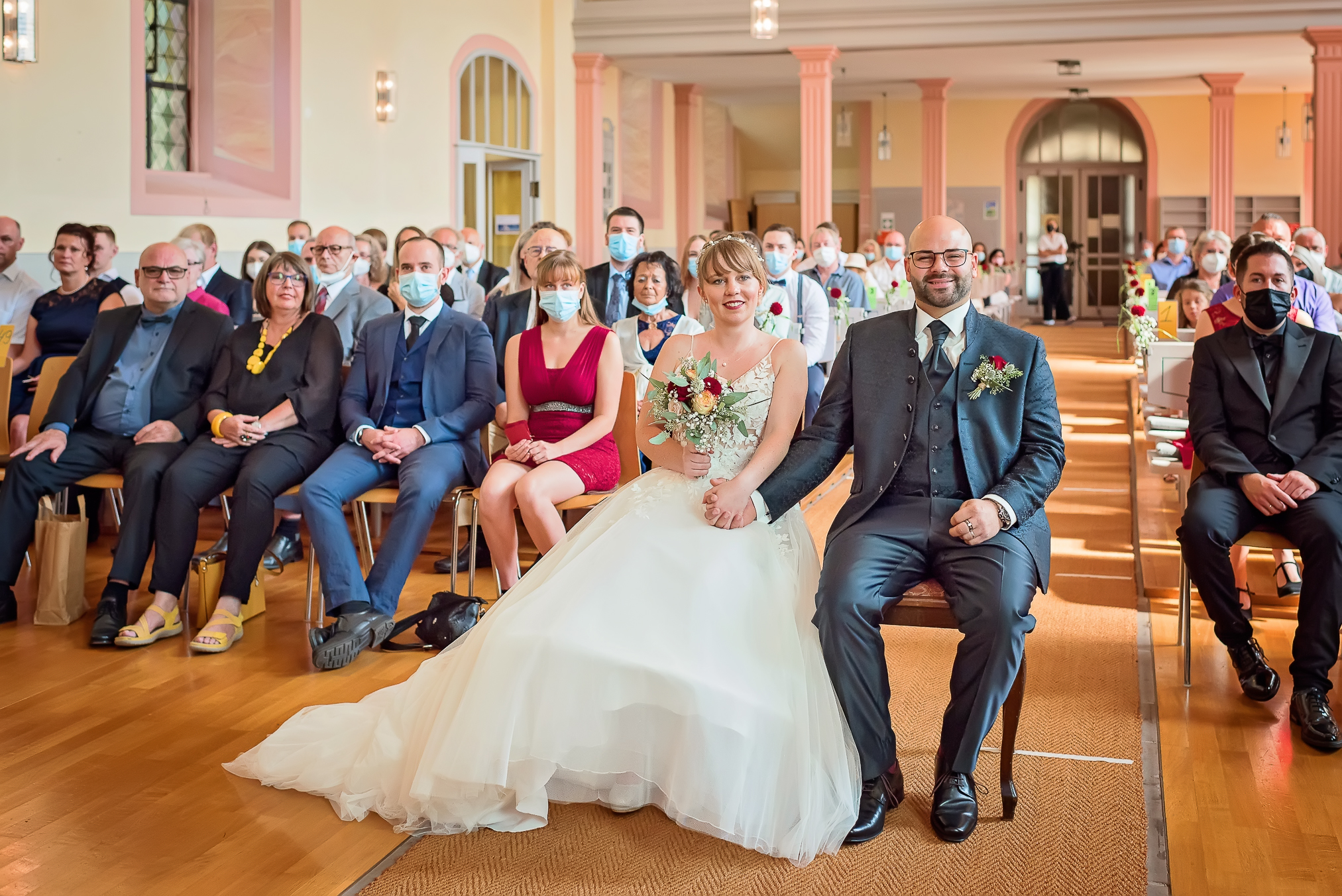 Foto eines Brautpaars, das während ihrer kirchlichen Trauung in der Ev. Martinskirche in Lahr mit verbundenen Händen zum Altar schaut und lacht. Im Hintergrund sind die Hochzeitsgäste zu sehen.