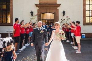 Foto des Brautpaars, das die Kirche nach ihrer Trauung verlässt und im Hintergrund von Volleyball-Freunden mit roten Blumen begrüßt wird.