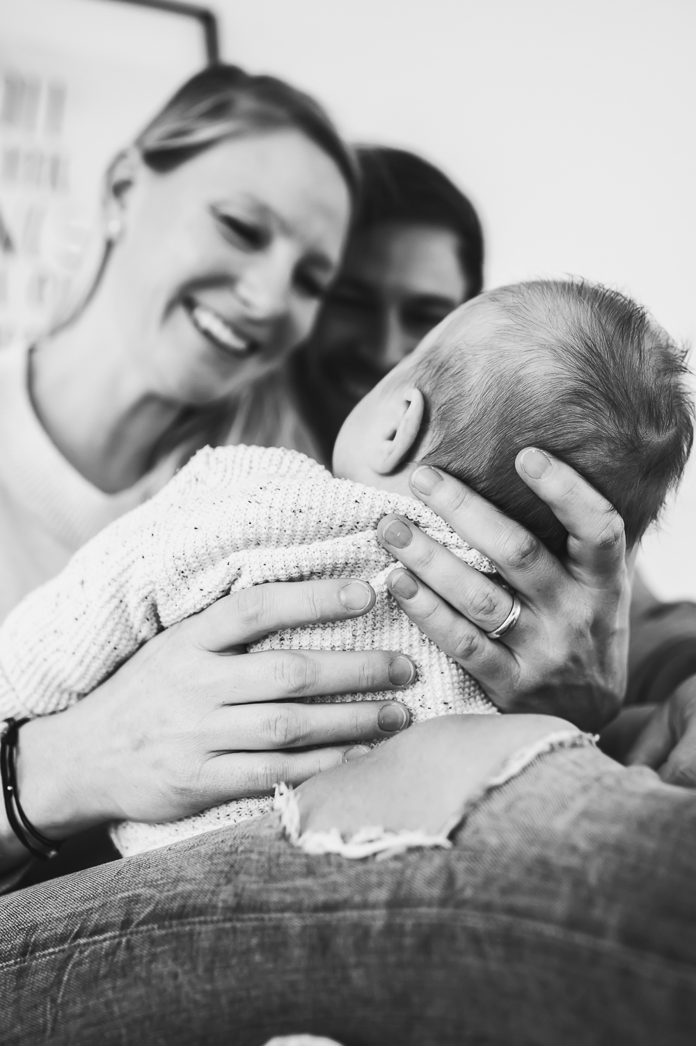 Ein berührendes Schwarz-Weiß-Porträt einer Familie mit einem neugeborenen Baby, das von der Mutter im Arm gehalten wird, während der Vater stolz auf das Baby schaut.