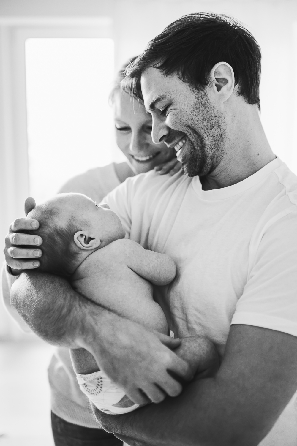 Ein berührendes Schwarz-Weiß-Porträt einer Familie, mit einer glücklichen Mutter und einem Vater, die ihr neugeborenes Baby liebevoll im Arm halten, während sie gemeinsam lachen.